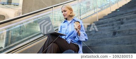 Image of stylish young modern woman, student doing homework, studying outdoors on campus stairs, sitting with laptop and coffee, drinking her cappuccino and connecting to public wifi Image of stylish young modern woman, student doing homework, studying outdoors on campus stairs, sitting with laptop and coffee, drinking her cappuccino and connecting to public wifi 109280053