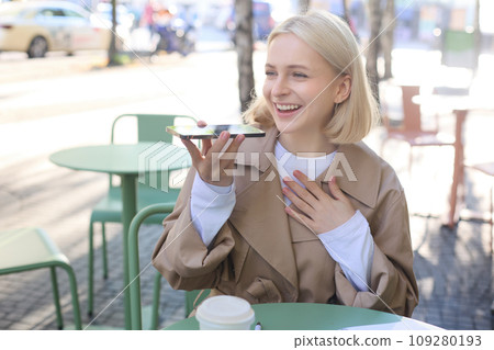 Lifestyle portrait of happy young woman, sitting in cafe and recording voice message, holding mobile phone near lips and talking into microphone, spending time in cafe, drinking coffee and chatting 109280193