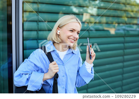 Close up portrait of blond smiling female model, college student in earphones, listening to music on street, holding smartphone, waiting for someone outdoors 109280229