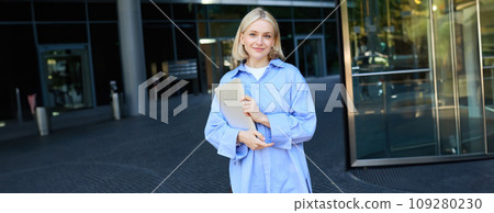 Image of young female employee, working woman standing near her office on street, holding pile of documents and smiling 109280230