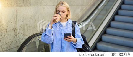 A happy, elegant businesswoman descending the escalator and holding takeaway coffee while reading important messages. A woman on the escalator with a phone A happy, elegant businesswoman descending the escalator and holding takeaway coffee while reading important messages. A woman on the escalator with a phone 109280239