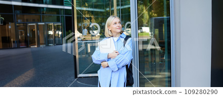 Image of young college girl, young woman with laptop, posing near university building, standing on street, smiling, concept of education and people Image of young college girl, young woman with laptop, posing near university building, standing on street, smiling, concept of education and people 109280290