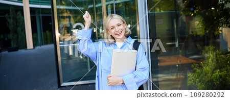 Portrait of happy, enthusiastic blond woman near office building, holding backpack and laptop, raising hands up and cheering, encouraging you, chanting and smiling Portrait of happy, enthusiastic blond woman near office building, holding backpack and laptop, raising hands up and cheering, encouraging you, chanting and smiling 109280292