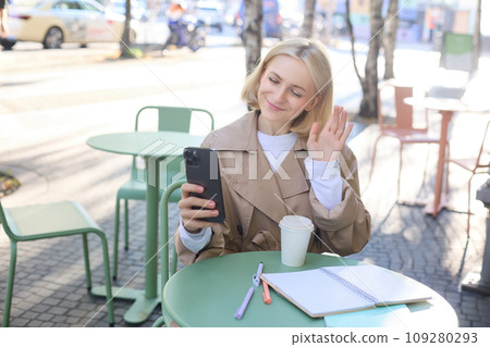 Portrait of young stylish woman in trench, sitting outdoors in city cafe, talking on video chat, using mobile phone to connect to online conversation, drinking coffee Portrait of young stylish woman in trench, sitting outdoors in city cafe, talking on video chat, using mobile phone to connect to online conversation, drinking coffee 109280293