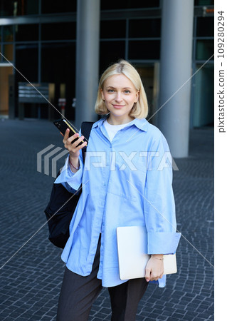 Vertical portrait of young office manager, woman in blue collar shirt and backpack, holding laptop and smartphone, waiting near business building on street, smiling at camera Vertical portrait of young office manager, woman in blue collar shirt and backpack, holding laptop and smartphone, waiting near business building on street, smiling at camera 109280294