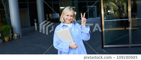 Young positive female model, student with notebooks and study material showing peace sign, standing near university, college campus, promo for new education scheme Young positive female model, student with notebooks and study material showing peace sign, standing near university, college campus, promo for new education scheme 109280985