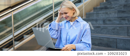Young woman, student in blue shirt, working on laptop, sitting outdoors on street stairs, working on project online, connects to public wifi, elearning 109281023