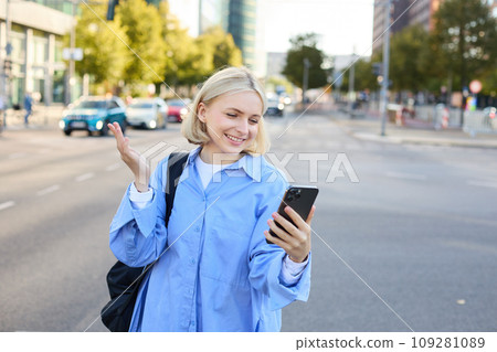Portrait of young office worker, woman walking along street, chatting on the phone, looking at smartphone and showing smth behind her, video chats, connects to online conversation via mobile app 109281089