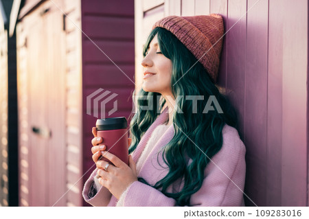 Stylish young smiling hipster woman with closed eyes wearing color hair, pink coat, hat holding reusable coffee cup leaning on beach hut and enjoying moment. Simple pleasures and personal fulfillment. 109283016