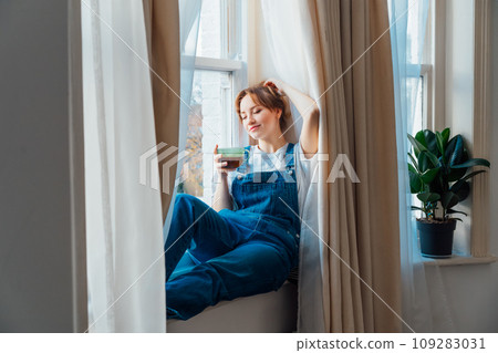 Free Time To Relax. Young woman looks out the window overlooking the city, sits on the windowsill at cozy home holds cup of hot tea drink. Happy calm female taking break for mental health wellbeing. Free Time To Relax. Young woman looks out the window overlooking the city, sits on the windowsill at cozy home holds cup of hot tea drink. Happy calm female taking break for mental health wellbeing. 109283031