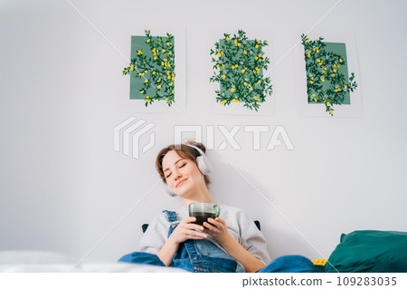 Woman with closed eyes listen music and enjoy cup of coffee or tea. Calm female spend free time at home enjoy favourite song with wireless modern headphones. Pastime weekend relax, no stress concept. Woman with closed eyes listen music and enjoy cup of coffee or tea. Calm female spend free time at home enjoy favourite song with wireless modern headphones. Pastime weekend relax, no stress concept. 109283035