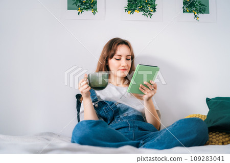 Young woman reading e-book on tablet reader in bed. Female relaxing with hot drink while reading an electronic book in cozy room. Mental health recovery, Personal growth. Hobby, Leisure time alone. Young woman reading e-book on tablet reader in bed. Female relaxing with hot drink while reading an electronic book in cozy room. Mental health recovery, Personal growth. Hobby, Leisure time alone. 109283041