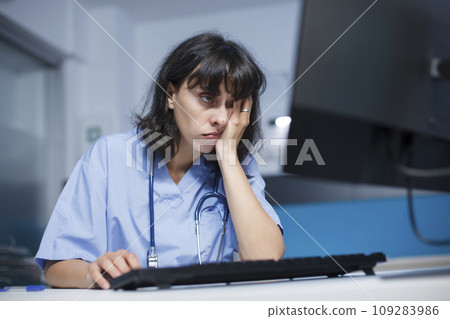 Close-up shot of a tired Caucasian woman with a stethoscope working on desktop PC in the clinic office. Selective focus of a worn out nurse in blue scrubs using a computer. Professional and detailed. Close-up shot of a tired Caucasian woman with a stethoscope working on desktop PC in the clinic office. Selective focus of a worn out nurse in blue scrubs using a computer. Professional and detailed. 109283986