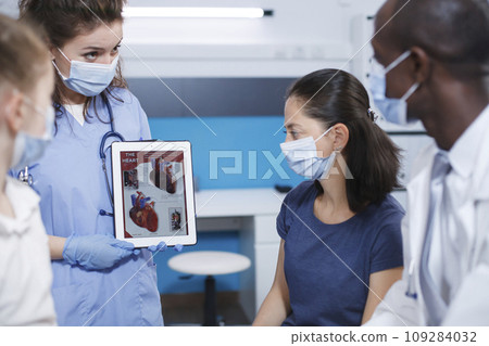 Detailed image of healthcare professionals using a tablet to explain to the female patients about the human heart. Nurse holds a device while the male doctor describes importance of medical checkups. 109284032