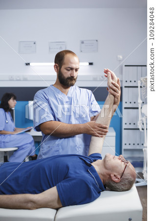 Male elderly patient receiving osteopathic care from a medical practitioner for arm pain. Caucasian retired old man aided by nurse in blue scrubs through physiotherapy exercise. Male elderly patient receiving osteopathic care from a medical practitioner for arm pain. Caucasian retired old man aided by nurse in blue scrubs through physiotherapy exercise. 109284048