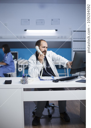 In medical office, male physician is using a computer for medical research and chatting on a wired phone. Caucasian man in lab coat using landline phone to speak with hospital receptionist. In medical office, male physician is using a computer for medical research and chatting on a wired phone. Caucasian man in lab coat using landline phone to speak with hospital receptionist. 109284092