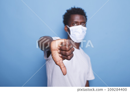 African american adult doing thumbs down gesture with right hand in studio. Black person showing sign while staring at camera and wearing face mask to protect from coronavirus pandemic. 109284400