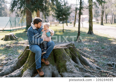 Dad with a little girl on his knees sits on a huge stump in a sunny spring park 109284695