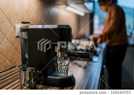 Empty glass stands on the stand of a carob coffee machine on a table against the background of a bartender Empty glass stands on the stand of a carob coffee machine on a table against the background of a bartender 109284703