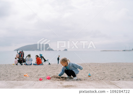 Little girl digs a hole in the sand while squatting against the background of mothers with children sitting on the shore 109284722