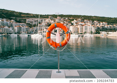Orange lifebuoy on a stand on a pier against the backdrop of a colorful resort town with moored yachts 109284783