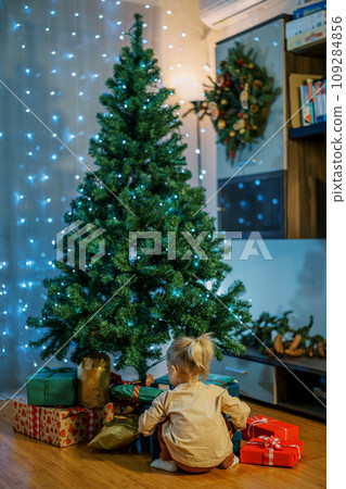 Little girl sits on the floor under the Christmas tree and looks at gifts. Back view 109284856