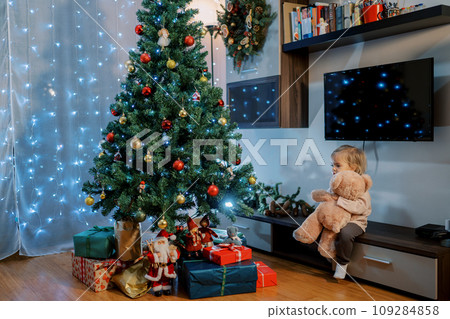 Little girl sits on a low locker, hugging a teddy bear near the Christmas tree 109284858