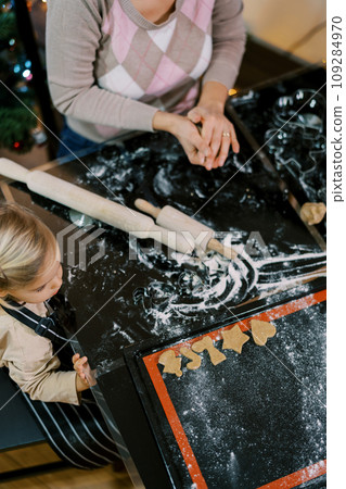 Little girl sits next to her mother at the table and looks at a tray of cut-out cookies. Top view Little girl sits next to her mother at the table and looks at a tray of cut-out cookies. Top view 109284970