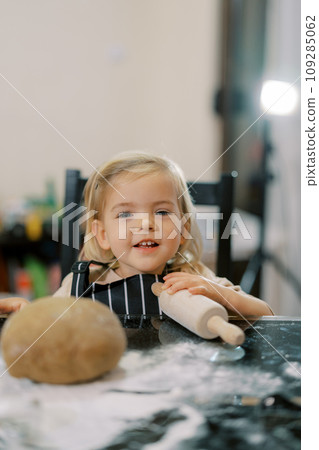Little smiling girl sits at a table with a rolling pin near the dough Little smiling girl sits at a table with a rolling pin near the dough 109285062