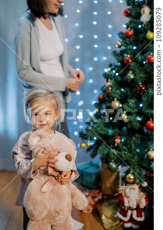 Smiling little girl hugging a teddy bear stands against the background of her mother decorating the Christmas tree. Cropped Smiling little girl hugging a teddy bear stands against the background of her mother decorating the Christmas tree. Cropped 109285079