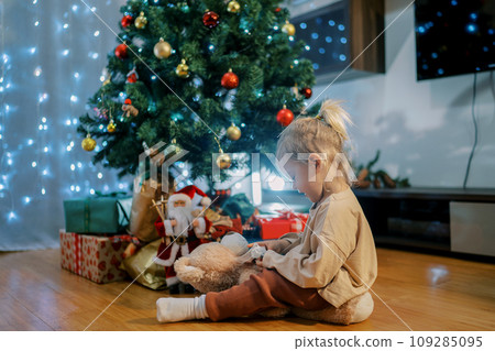 Little girl examines a bow in her hands while sitting on a teddy bear near a shining Christmas tree 109285095