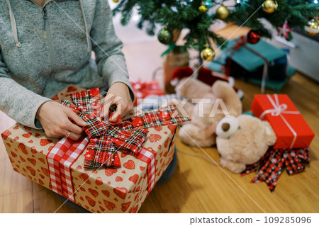 Girl unties a bow on a colorful gift box sitting near the Christmas tree. Cropped. Faceless 109285096