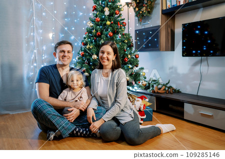Mom, dad and little girl are sitting on the floor near the decorated Christmas tree Mom, dad and little girl are sitting on the floor near the decorated Christmas tree 109285146