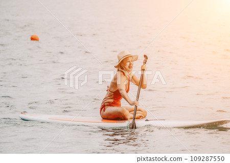 Woman in red bikini on sup board. Happy lady with blond hair in red bathing suit chilling and sunbathing by turquoise sea ocean on sunset. Holiday, vacation and recreational concept. Woman in red bikini on sup board. Happy lady with blond hair in red bathing suit chilling and sunbathing by turquoise sea ocean on sunset. Holiday, vacation and recreational concept. 109287550