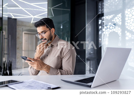 Businessman thinking while sitting inside office at table, man is concentrating and seriously reading online using an application on phone, the young boss is holding a smartphone in his hands. 109293827