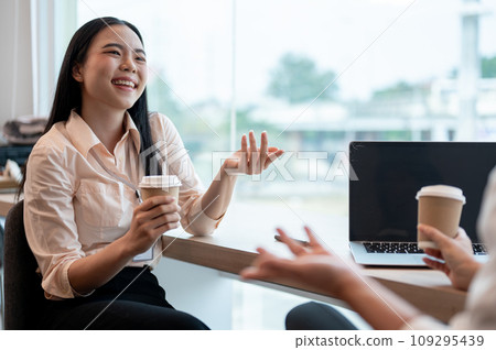 A cheerful Asian businesswoman is talking with her colleague during a coffee break in the office. A cheerful Asian businesswoman is talking with her colleague during a coffee break in the office. 109295439