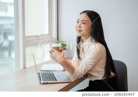 An attractive Asian businesswoman is daydreaming while enjoying her coffee in the office. 109295447