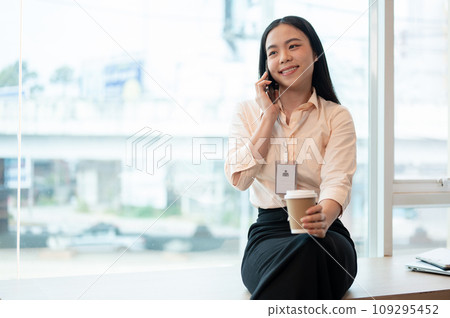 A beautiful Asian businesswoman is talking on the phone while having her coffee in the office. A beautiful Asian businesswoman is talking on the phone while having her coffee in the office. 109295452