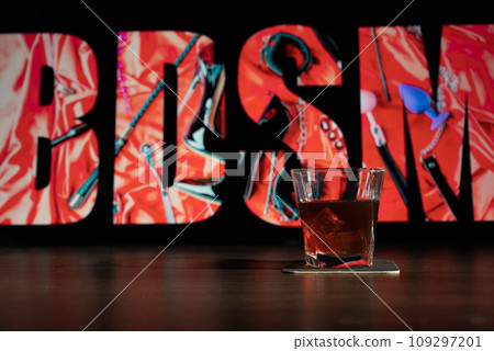 Glass of whiskey on the bar in front of the blur image Creative BDSM lettering on a black background. Set of sexy erotic toys Glass of whiskey on the bar in front of the blur image Creative BDSM lettering on a black background. Set of sexy erotic toys 109297201