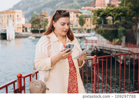 Hipster girl with two tickets browsing Internet. Cruise ship vacation concept. Varenna, Italy. Lake Como. Phone Communication. Female holding phone and booking hotel Hipster girl with two tickets browsing Internet. Cruise ship vacation concept. Varenna, Italy. Lake Como. Phone Communication. Female holding phone and booking hotel 109297788