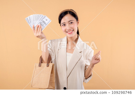 portrait of happy successful confident young asian business woman wearing white jacket holding cash money dollars and eco bag standing over beige background. Eco friendly shopping concept. portrait of happy successful confident young asian business woman wearing white jacket holding cash money dollars and eco bag standing over beige background. Eco friendly shopping concept. 109298746