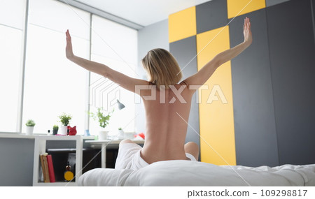 Rear view of naked woman looking out the window and raising her hands up while sitting on bed 109298817