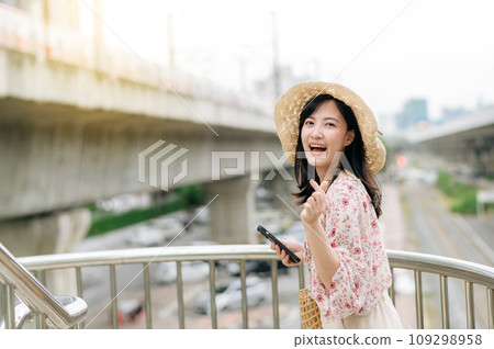 young asian woman traveler with weaving basket using mobile phone and standing on overpass with railway background. Journey trip lifestyle, world travel explorer or Asia summer tourism concept. 109298958