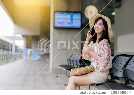 young asian woman traveler with weaving basket using a mobile phone and waiting for train in train station. Journey trip lifestyle, world travel explorer or Asia summer tourism concept. 109298964