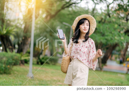 Portrait of asian young woman traveler with weaving hat and basket using mobile phone on green park nature background. Journey trip lifestyle, world travel explorer or Asia summer tourism concept. 109298990