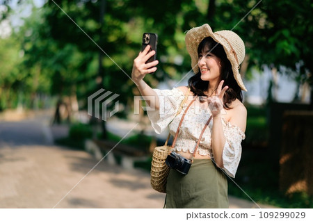 Portrait of asian young woman traveler with weaving hat, basket, mobile phone and camera on green public park background. Journey trip lifestyle, world travel explorer or Asia summer tourism concept. Portrait of asian young woman traveler with weaving hat, basket, mobile phone and camera on green public park background. Journey trip lifestyle, world travel explorer or Asia summer tourism concept. 109299029