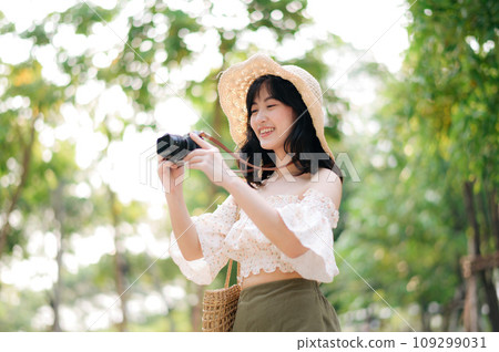 Portrait of asian young woman traveler with weaving hat and basket and a camera on green public park nature background. Journey trip lifestyle, world travel explorer or Asia summer tourism concept. Portrait of asian young woman traveler with weaving hat and basket and a camera on green public park nature background. Journey trip lifestyle, world travel explorer or Asia summer tourism concept. 109299031