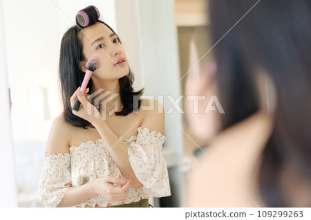 Beautiful young Asian woman applying makeup in front of the mirror. Lifestyle people concept. Advertisement for skin cream, anti-wrinkle cream, baby face Beautiful young Asian woman applying makeup in front of the mirror. Lifestyle people concept. Advertisement for skin cream, anti-wrinkle cream, baby face 109299263