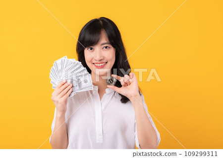 Portrait beautiful young asian woman enterpriser happy smile wearing white shirt and red plants holding cash dollar money and crypto digital currency isolation on yellow background. Wealth concept. 109299311