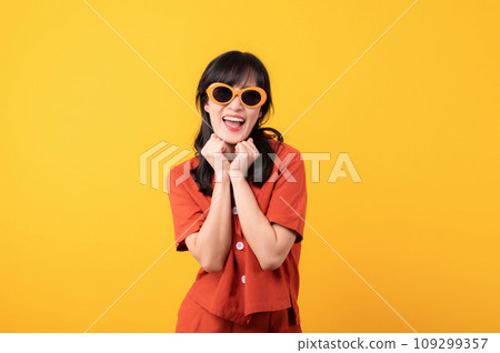 Portrait young beautiful asian woman cute and shy dressed in orange clothes and sunglasses touching cheeks showing shy emotional feeling isolated on yellow studio background. Portrait young beautiful asian woman cute and shy dressed in orange clothes and sunglasses touching cheeks showing shy emotional feeling isolated on yellow studio background. 109299357